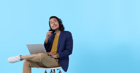 Obraz premium Young asian businessman thinking. While her using laptop sitting on chair and looking isolate on blue background.