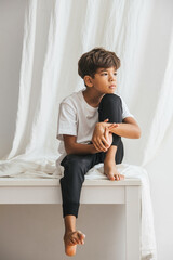 Thoughtful bored and sad indian boy sitting on a table barefoot, looking nowhere. Against white curtain.