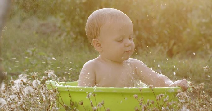 A cute little baby boy plays in a bowl of water in a summer garden on a sunny day