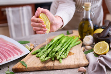 Woman squeezing lime juice onto Green Asparagus on wooden cutting board and slice bacon at kitchen table