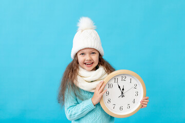 cute little girl child in winter clothes hat and sweater with a watch in her hands on a blue...
