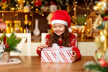 cute little girl child in winter clothes Santa Claus hat and red sweater rejoices at the gift and...