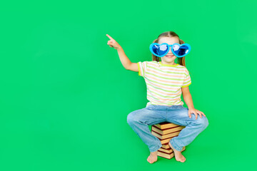 a girl child sits on a green background on books in blue large funny glasses and points her finger up or to the side, space for text