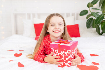 the concept of Valentine's day, a cute child girl is sitting on the bed at home in red pajamas and holding a gift in her hands and smiling or laughing with happiness, congratulating on the holiday
