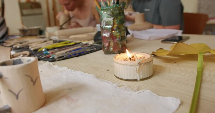 a candle burns on the table in the workshop with tools on the background of girls sculpting dishes from clay. production of handmade sculptures. close-up