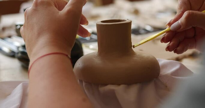 female hands make patterns on pottery with a plasma yellow spatula at the table in the workshop against the background of a window and a camera. general plan bokeh