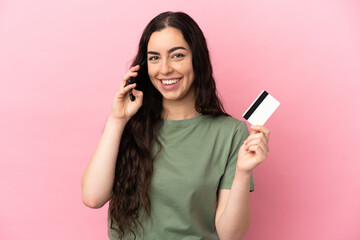 Young caucasian woman isolated on pink background keeping a conversation with the mobile phone and holding a credit card