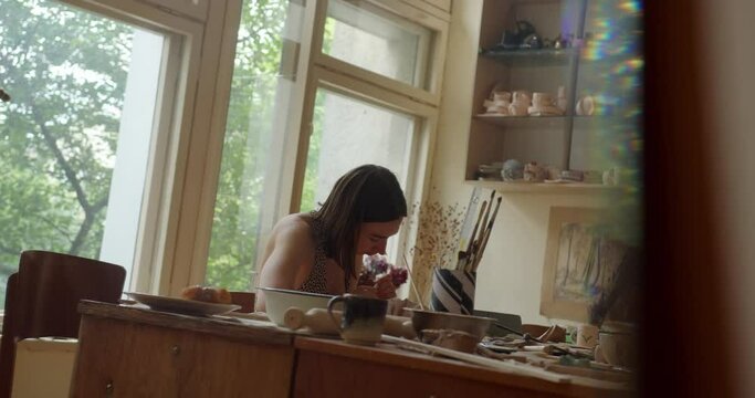 a girl sculptor makes figures from clay in the workshop against the background of dishes on the shelves and a window. the girl sculpts ceramics at the table. view from a glass prism.