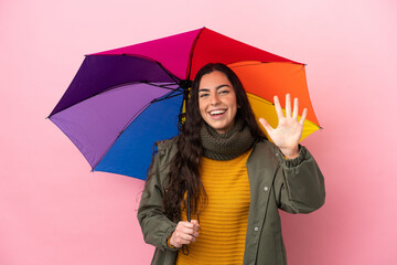 Young woman holding an umbrella isolated on pink background counting five with fingers