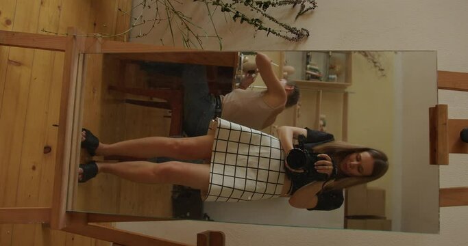 a beautiful girl in a short checkered skirt takes a picture of herself on camera in a full-length mirror in a pottery workshop, in the background girls are sculpting cups from clay. vertical video