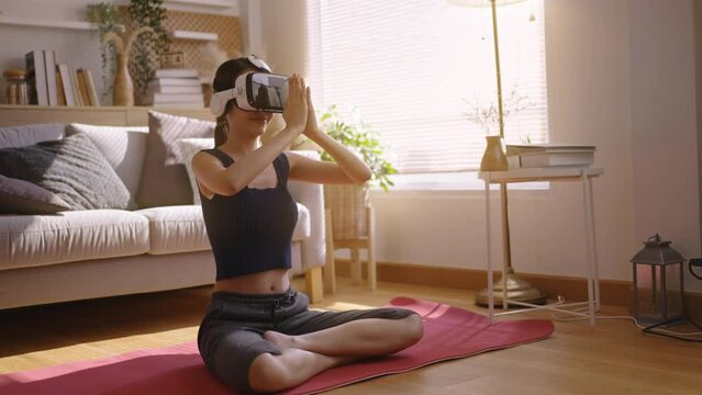 Asian Female woman wearing virtual reality glasses meditating in lotus position at home,yoga namaste enjoying simulation and relax in metaverse world.AR,VR Virtual Future technology concept