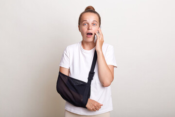 Woman wearing casual t shirt posing isolated over white background, girl has broken arm talks via smartphone looks at camera with shocked expression. People health problems and technology.