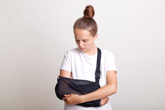 Indoor shot of woman with bun hairstyle with broken arm in splint suffers from hand injury, insurance and health care, girl wearing casual t shirt posing isolated over white background.