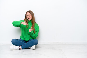 Young caucasian woman sitting on the floor isolated on white background pointing to the front and smiling