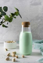 Pistachio milk in a transparent bottle with a cork on a light table. Pistachios in a white vase in the background. Light green napkin on the table. Light background