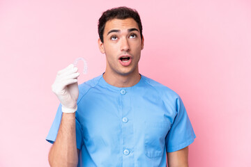 Dentist caucasian man holding invisible braces isolated on pink background looking up and with surprised expression