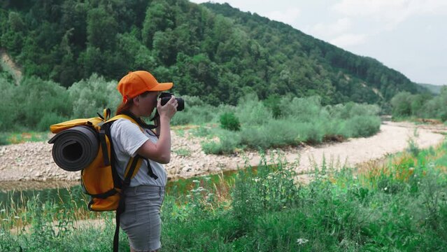 Hiking with camera, orange backpack and cap. A tourist girl takes pictures of nature during a trip. A young woman takes pictures on a DSLR camera. A trip to rest in nature in the forest and mountains.