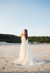 Fototapeta na wymiar Beautiful bride in long beautiful wedding dress. Beautiful girl in wedding dress posing in front of desert, sand, canyon with a bouquet in her hands