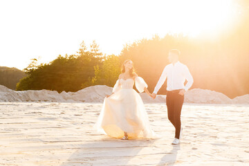 Fototapeta na wymiar Newlyweds in love, holding hands, run along a sandy quarry at sunset.