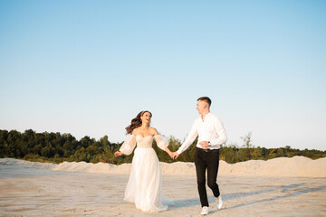 Luxury newlyweds are running on the sand against the background of the forest, holding hands and looking at the sun