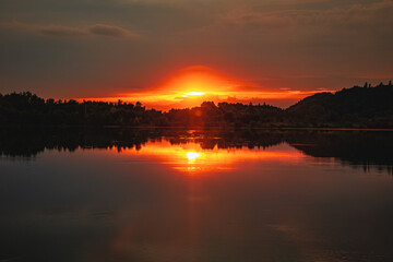 Epic red and gold clouds over forest lake at sunset. Dramatic cloud cover. Symmetrical reflections on the water, natural mirror. idyllic landscape.