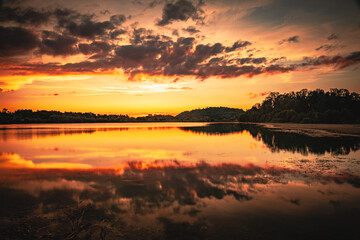 Naklejka na ściany i meble Epic red and gold clouds over forest lake at sunset. Dramatic cloud cover. Symmetrical reflections on the water, natural mirror. idyllic landscape.