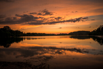 Naklejka na ściany i meble Epic red and gold clouds over forest lake at sunset. Dramatic cloud cover. Symmetrical reflections on the water, natural mirror. idyllic landscape.