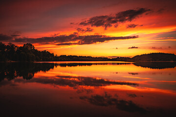 Fototapeta na wymiar Epic red and gold clouds over forest lake at sunset. Dramatic cloud cover. Symmetrical reflections on the water, natural mirror. idyllic landscape.