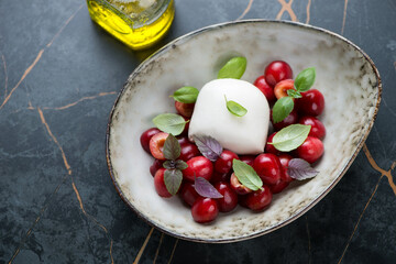 Beige bowl with mozzarella cheese, cherries and fresh basil leaves, elevated view on a dark-olive marble background, studio shot