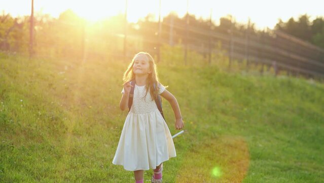 Elementary schoolgirl runs on green grass after classes at school. Schoolkid wearing blue backpack and white dress runs skipping at back sunset light