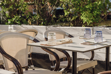 Empty table and chairs on the summer terrace near a small cafe. The restaurant is open and waiting for guests. A laid cafeteria table outside. There are glasses, napkins and cutlery on the table.