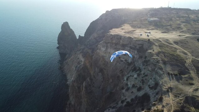 Aerial drone view of a man flying a white and blue paraglider over a hill and trees to the sea waves near the rocks. Active paraglider flight over the seascape with clear skies at suset. Extreme sport