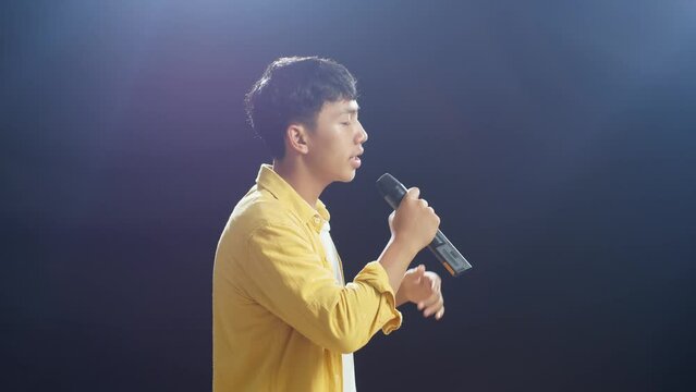 Side View Of Young Asian Boy Holding A Microphone And Rapping On The Black Background
