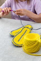 On table is ball of yellow knitted yarn with ready-made soles for slippers. In blurry background, women's hands are knitting. Selective focus. Images for websites about knitting, hobbies