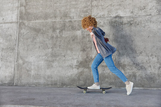 Energetic hipster girl rides skateboard dressed in casual clothes enjoys spare time poses against grey concrete wall with blank space for your promotional content. People pastime and recreation