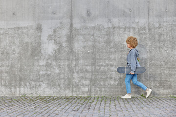 Fototapeta na wymiar Active teenage girl with curly hair dressed in hoodie jeans and sneakers carries longboard enjoys active rest strolls outdoors against grey concrete wall empty space for your promotional content