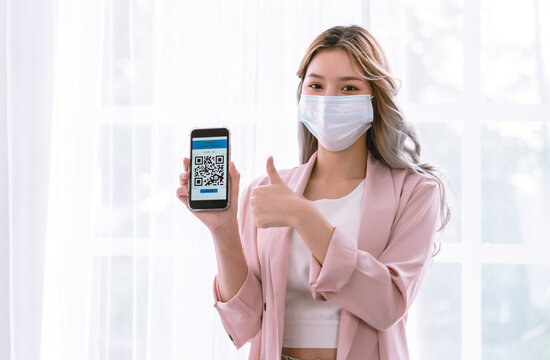 Portrait of beautiful Asian woman with face mask show health passport for travel during covid-19 pandemic and show thumps up with positive emotions. Technology QR code, pandemic prevention concept.