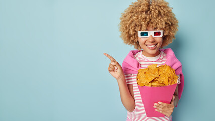 Happy curly haired young woman invites you to cinema points away on blank space advertises intersting film to watch holds paper bucket of crisps wears 3d glasses poses against blue background.