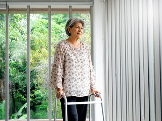 Asian senior woman, white hair standing with walking frame and looking out the glass window,...