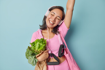 Positive Asian sportswoman dances carefree keeps arm raised up listens music via headphones dressed in spotswear carries expander embraces paper bag with vegetables moves against blue background