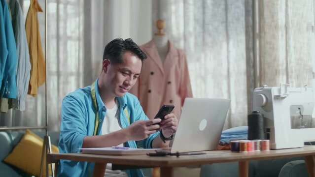 Asian Male Designer With Sewing Machine Looking At Smartphone While Designing Clothes On Laptop Computer In The Studio
