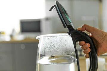 A hand opens the lid of an electric kettle. A modern electric transparent kettle on a wooden table in the kitchen. A kettle for boiling water and making tea. Home appliances for making hot drinks.