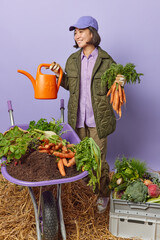 Happy Asian woman wears cap and jacket holds watering can and bunch of carrots poses around harvested vegetables in domestic garden grows healthy homegrown vegetarian food. Seasonal autumn harvest