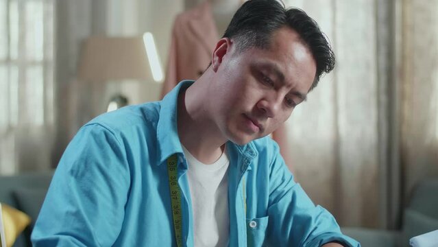 Close Up Of Asian Male Designer With Sewing Machine Designing Clothes On Paper While Working With Laptop Computer In The Studio
