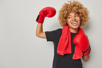 Waist up shot of curly haired sportswoman wears red boxing gloves shows strong biceps dressed in black t shirt and towel around neck poses against grey background empty space for your promo. - obrazy, fototapety, plakaty