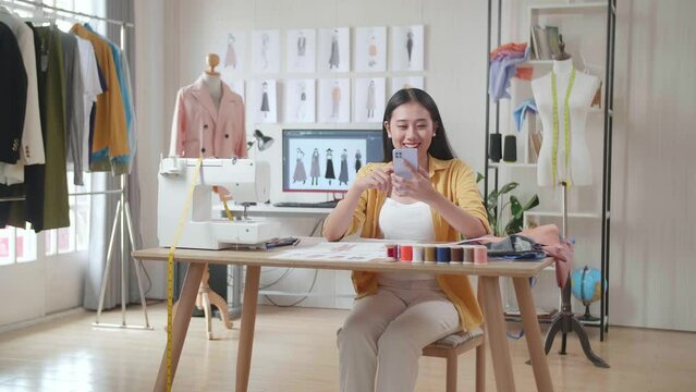Smiling Asian Female Designer With Sewing Machine Looking At The Paper In Hand And Comparing It To The Pictures On Smartphone While Designing Clothes In The Studio
