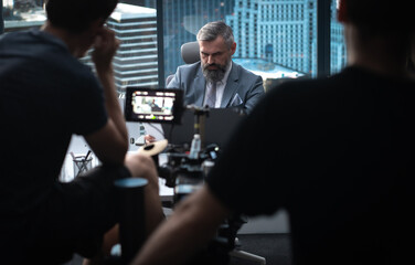 Behind the scenes - Film crew shooting a movie or a corporate video. Adult grey-haired Caucasian CEO businessman actor sitting at in the office located in a skyscraper
