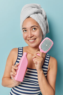 Beautiful young Asian woman smiles happily concentrated above recommends shapoo for hair care holds comb wears striped t shirt and bath towel wrapped over head isolated over blue background.