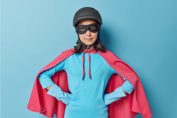 Self confident woman dressed in superhero costume keeps hands on waist wears protective helmet...
