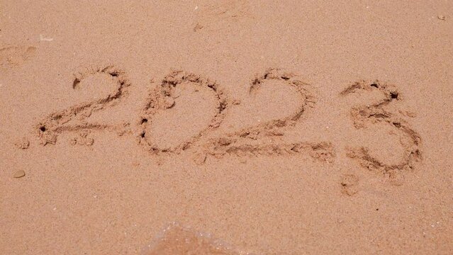 New year 2023 handwritten is appearing after washing away 2022 y on sandy beach.